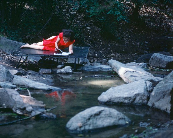 The Woman in Red, on a bed jutting into the water, skims an accordion-folded album of photos, performance Rivers First Draft by Lorraine O’Grady.